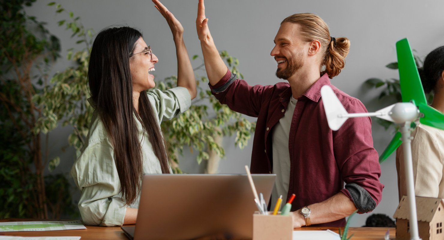 Two colleagues celebrate making their website accessible with a high-five in a sustainable, eco-friendly office setting.